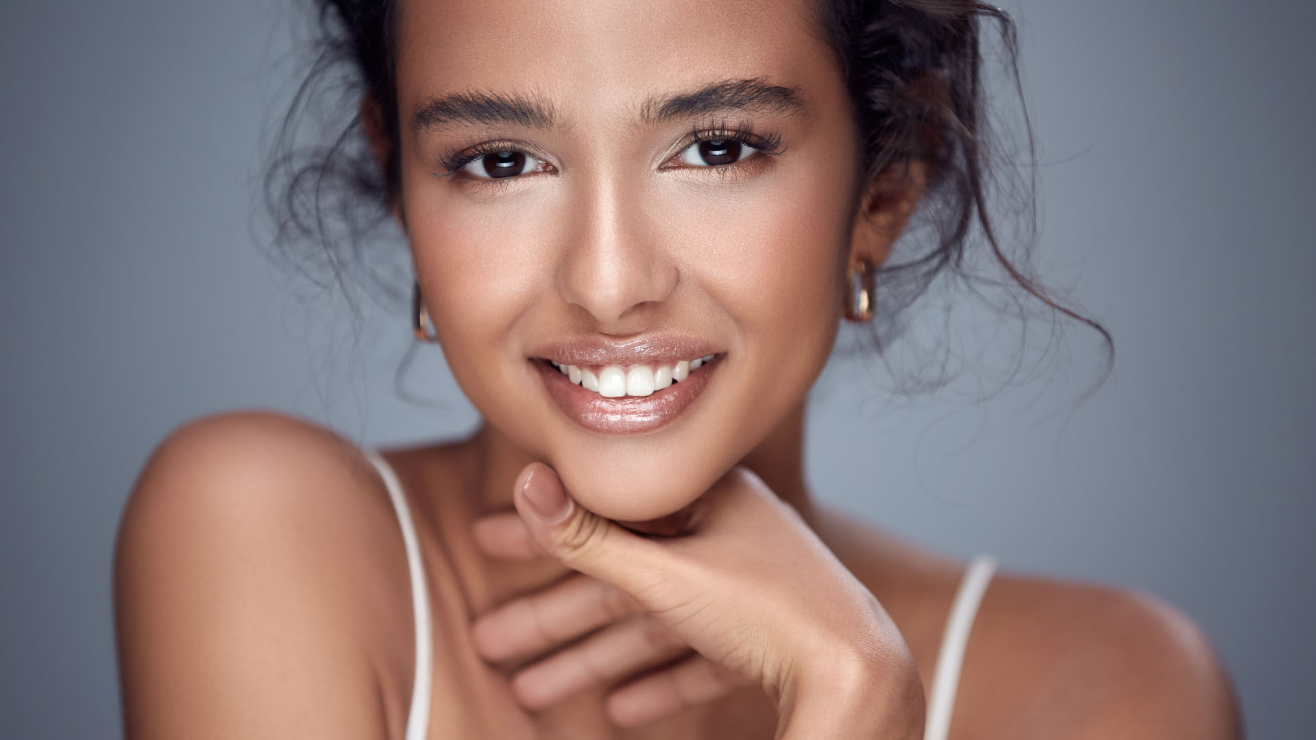 Young woman with clear skin smiling warmly, resting her chin on her hand against a grey background